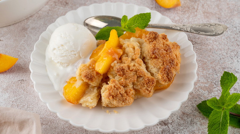A piece of peach cobbler on a scalloped white plate with vanilla ice cream and mint leaves