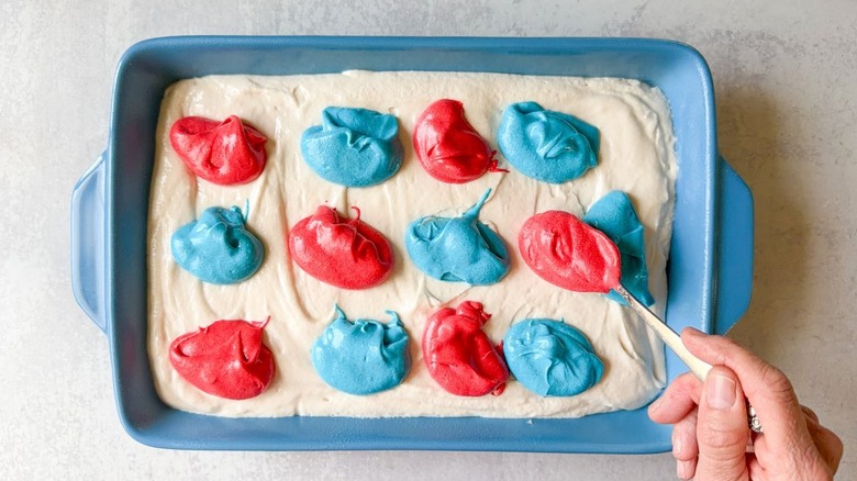Dropping spoonfuls of red and blue cake batter on top of vanilla cake batter in blue baking dish