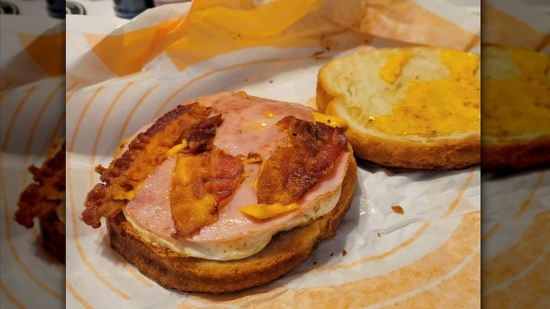 A Jack in the Box Ultimate Breakfast Sandwich rests with its top off beside the fillings on a paper wrapper