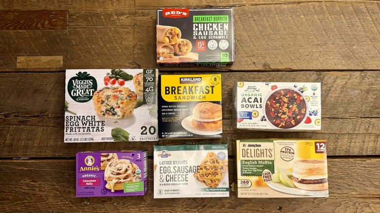 An assortment of Costco frozen breakfast items on a wooden table, including Red's Chicken Sausage and Egg Scramble Breakfast Burrito, Veggies Made Great Spinach and Egg White Frittatas, Kirkland Breakfast Sandwich, Organic Acai Bowls, Annie's Cinnamon Rolls, Lattice d'Lights Egg Sausage and Cheese Pockets, and Jimmy Deans English Muffin Delights