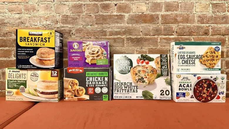 An assortment of Costco frozen breakfast items against a brick wall, including Red's Chicken Sausage and Egg Scramble Breakfast Burrito, Veggies Made Great Spinach and Egg White Frittatas, Kirkland Breakfast Sandwich, Organic Acai Bowls, Annie's Cinnamon Rolls, Lattice d'Lights Egg Sausage and Cheese Pockets, and Jimmy Deans English Muffin Delights