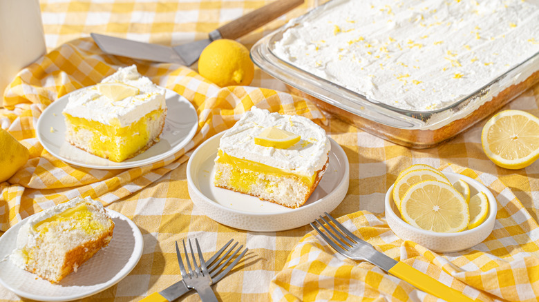 Lemon cake with white frosting surrounded by slices of cake and lemons on a sunny picnic blanket.