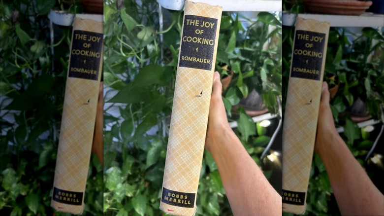 A hand holds up Irma Rombauer's "The Joy of Cooking" in an outdoor setting.