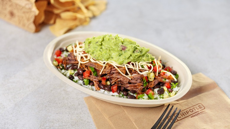 A burrito bowl from Chipotle sits on a white table with some spilled tortilla chips.