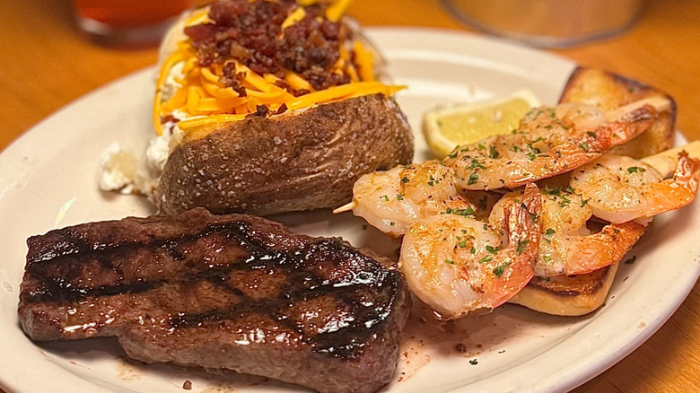 A white plate contains a steak, baked potato, and shrimp skewers