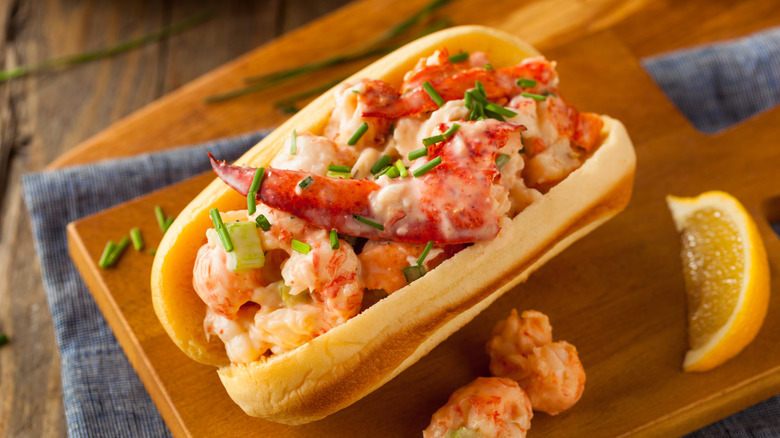 A lobster roll on a brioche bun sits on a wooden serving board with a lemon slice