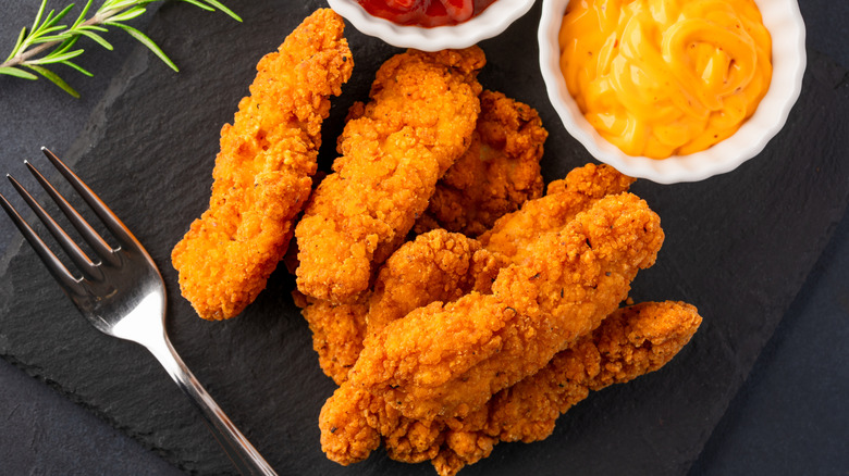 A pile of chicken tenders lie on a piece of black stale next to condiments