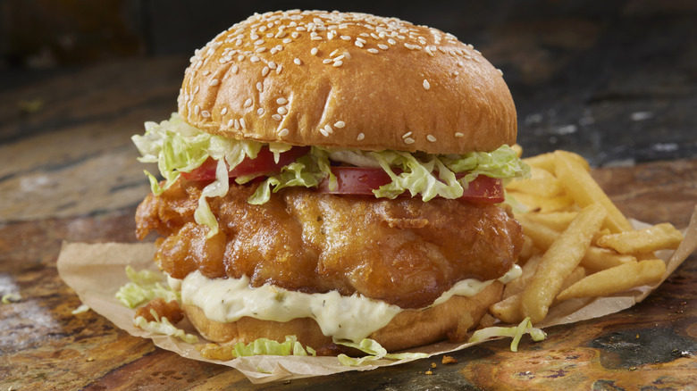 A fish sandwich with lettuce, tomato, and tartar sauce sits next to a pile of fries