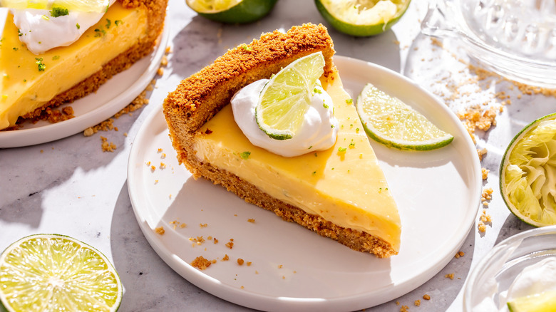 A slice of key lime pie surrounded by limes and graham cracker crumbs