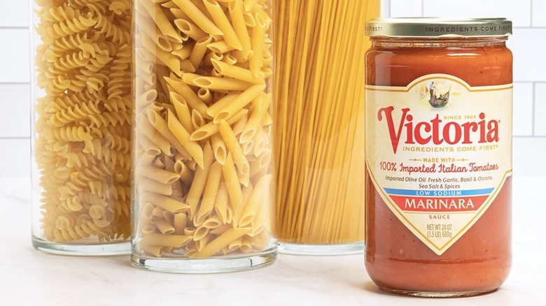 Jar of Victoria marinara beside containers of pasta before a white tile backsplash