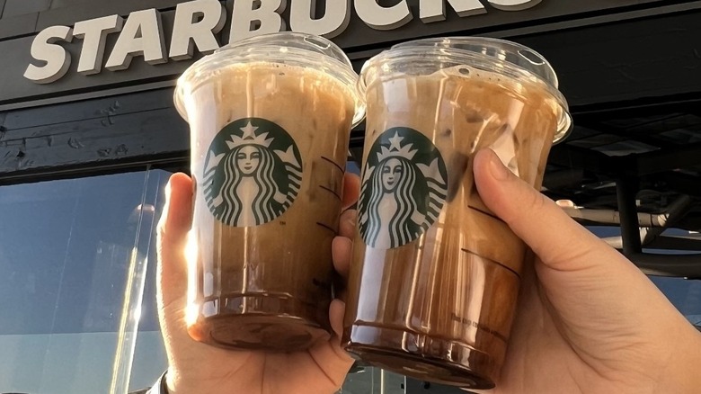 Two Starbucks iced shaken espressos are held up in front of a Starbucks store
