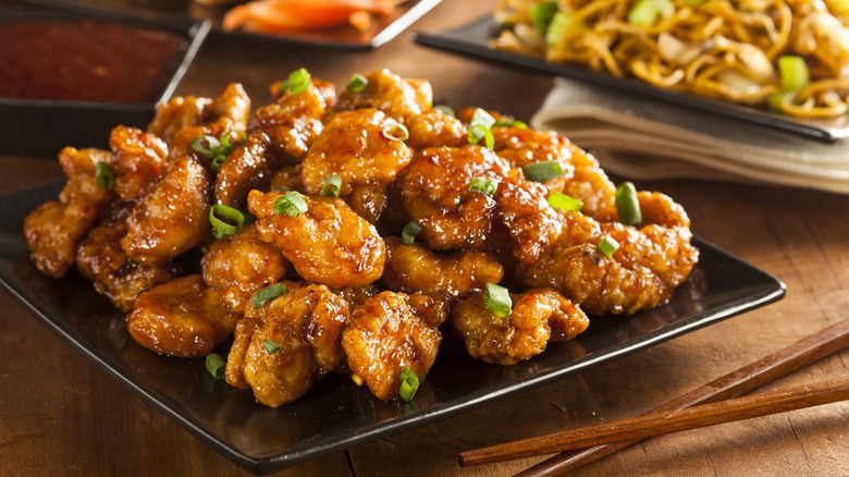 A plate of orange chicken with scallions sits on a wooden table next to a pair of chopsticks