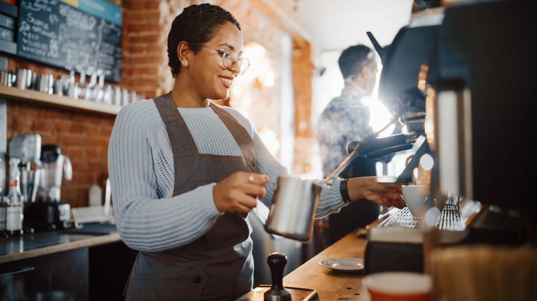 A smiling barista steams milk in a silver pitcher