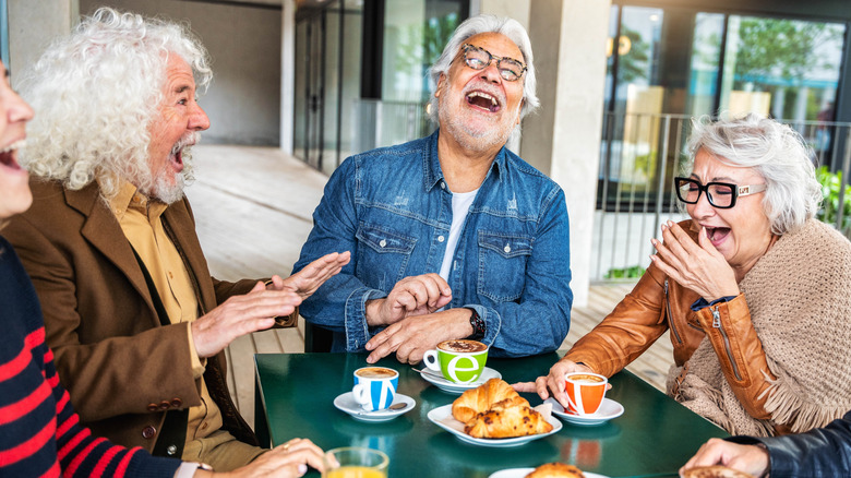 A group of senior citizens laugh together over coffee and croissants