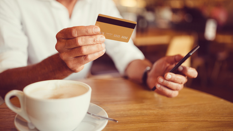 An older man holds up a credit card by a cup of coffee in a coffee shop