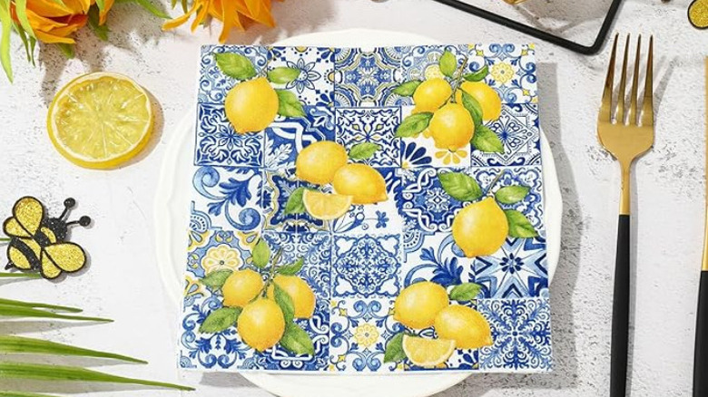 Greek tile inspired napkin design with pictures of lemons on a plate next to a brass and black fork, lemon wedge, and flowers