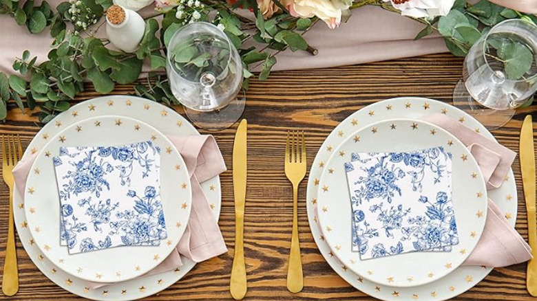 Blue flower design on white napkins on plates with gold cutlery and empty wine glasses