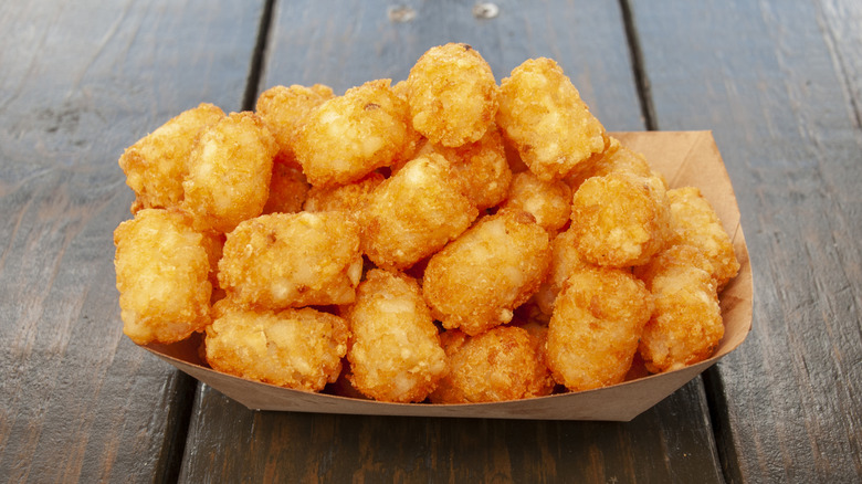 A large order of tater tots sits in a paper boat on a wooden table