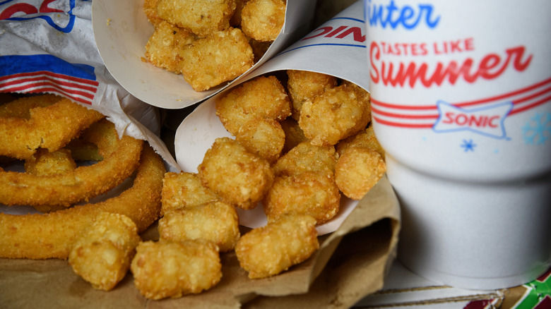 Two order of Sonic tater tots sit next to a bag of onion rings and a fountain drink