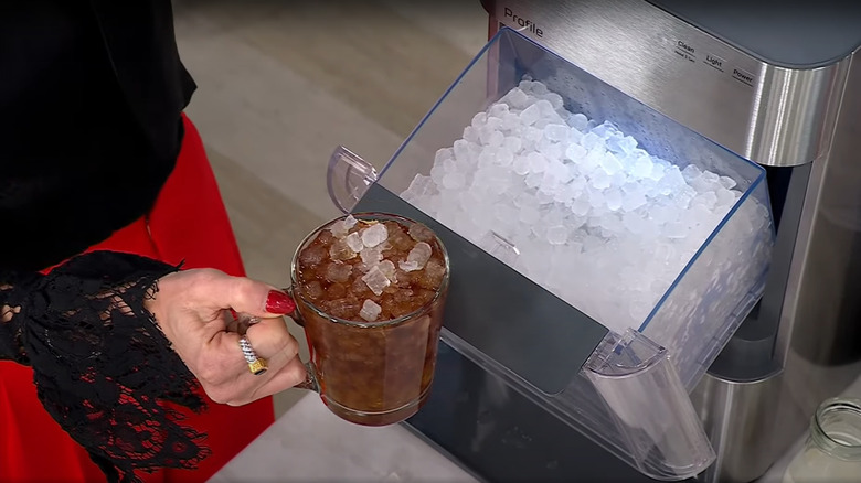 Someone holds a mug of iced coffee, filled with nugget ice, next to the GE nugget ice maker