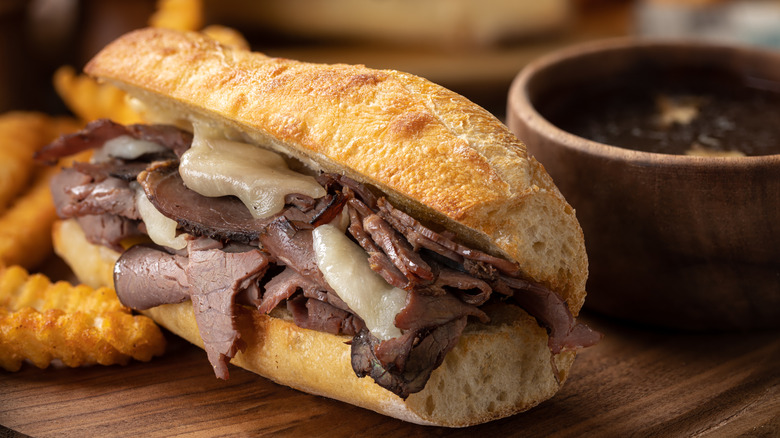 A French dip sandwich with cheese sits next to fries and au jus on a wooden serving board