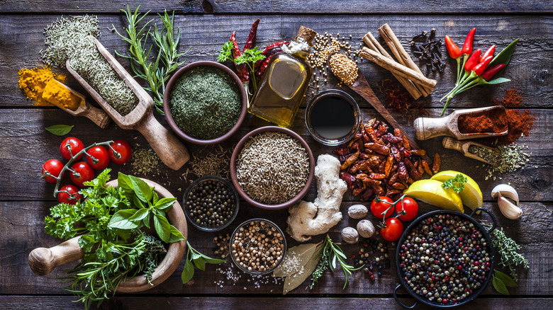A selection of dried and fresh herbs and spices rest next to fresh ingredients like tomatoes, ginger root, and dried peppers