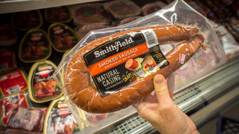 A person holds a pack of Smithfield smoked sausage by a grocery store display case