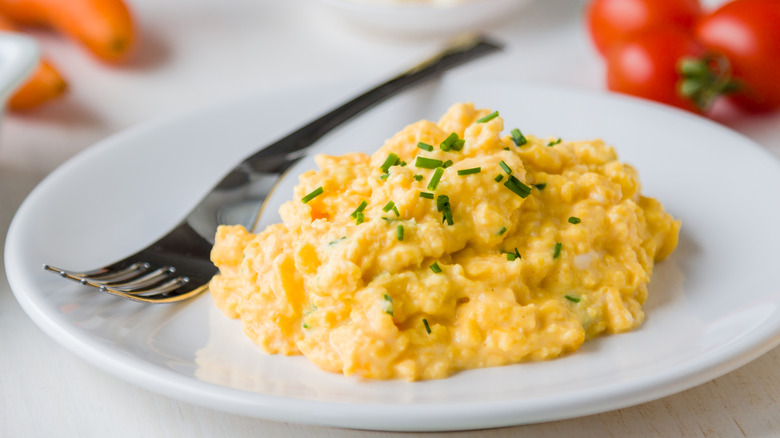 A pile of scrambled eggs with chive garnish sits on a white plate next to a fork