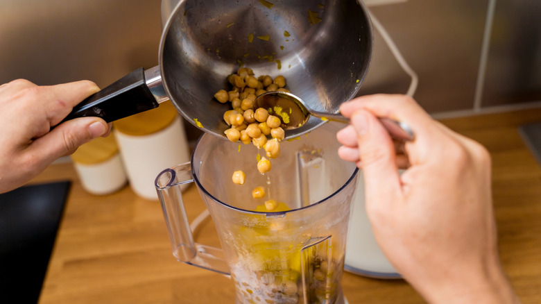 Hands using spoon to add chickpeas to blender canister