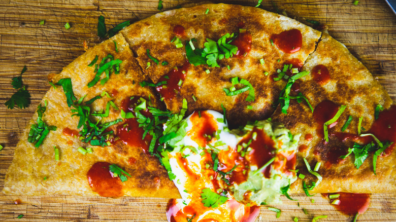Overhead view of a crispy quesadilla topped with fresh herbs, hot sauce, and crema