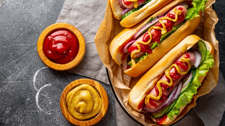 Three hot dogs with toppings next to bowl of ketchup and mustard