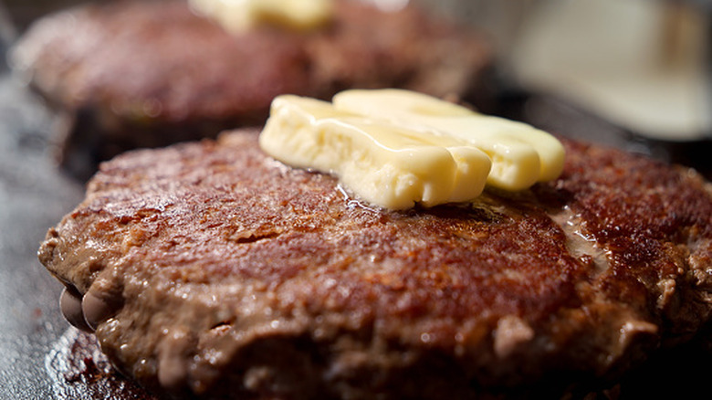 Two pats of butter melt on a burger patty cooking on a flat top.