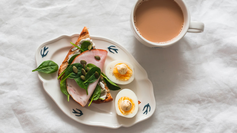 A boiled egg sliced in half and topped with butter and seasonings sits on a plate also containing bread topped with lunchmeat, spinach, and pumpkin seeds next to a cup of coffee