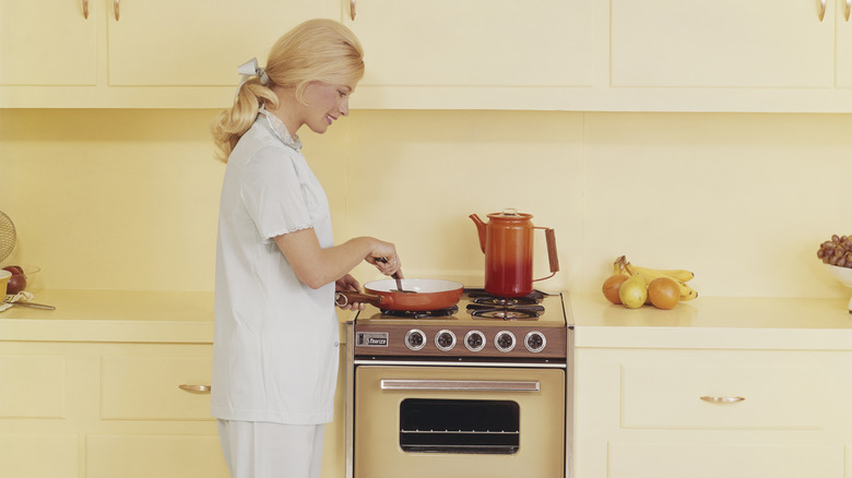 A smiling blonde woman stands at a skillet over a kitchen stove in a 1970s-era kitchen