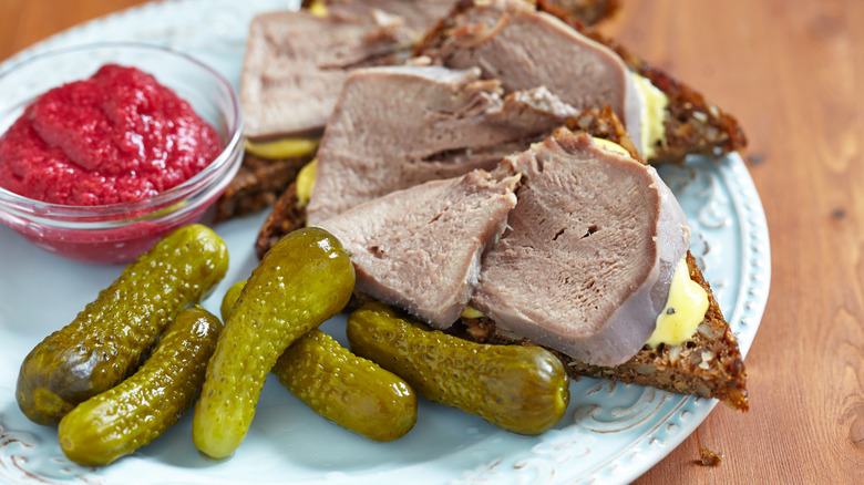 Open-faced beef tongue sandwiches with pickles and a red sauce on the side on a white plate