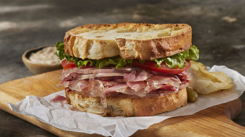 Head cheese sandwich with tomatoes and lettuce on thick bread with chips on butcher paper on a wooden cutting board