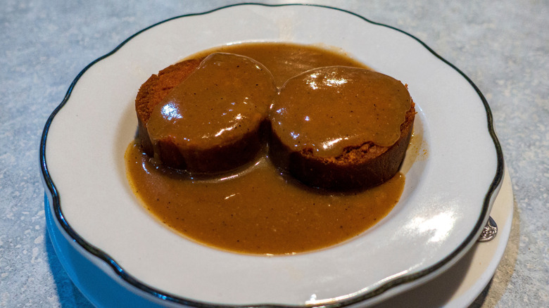 Two slices of kishke with a brown gravy on a white plate with a black painted rim
