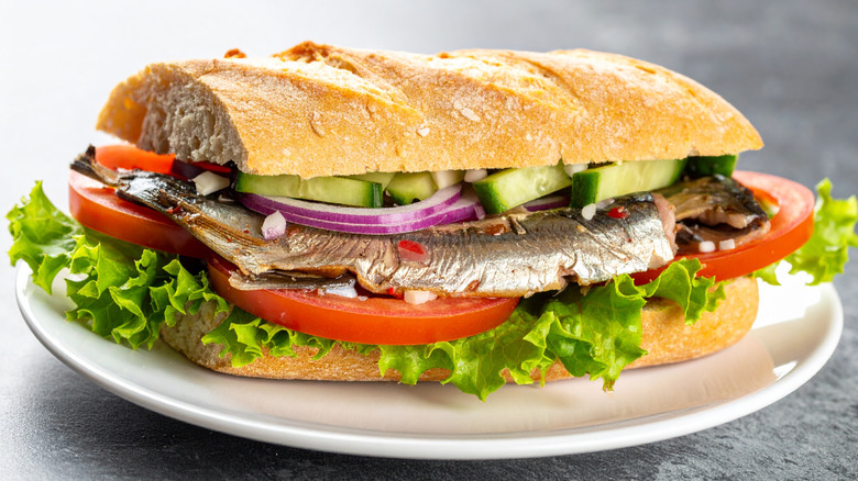 sardine sandwich with cucumbers, red onions, tomatoes, and lettuce on a white plate