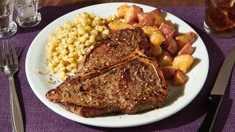 A T-bone steak dinner from Denny's on a white plate, including corn and country potatoes, sits on a purple placemat