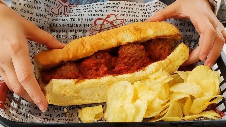 A person picks up a large meatball sub from a paper lined tray with potato chips in Goodcents