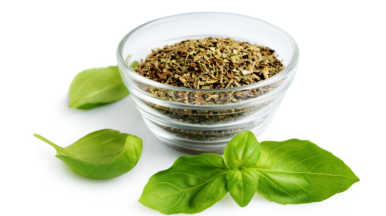A small bowl of dried Italian seasonings next to fresh leaves of basil