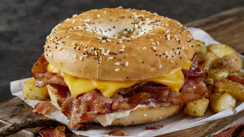 A fast food everything bagel with cheese and bacon sits beside fries on a wooden serving board