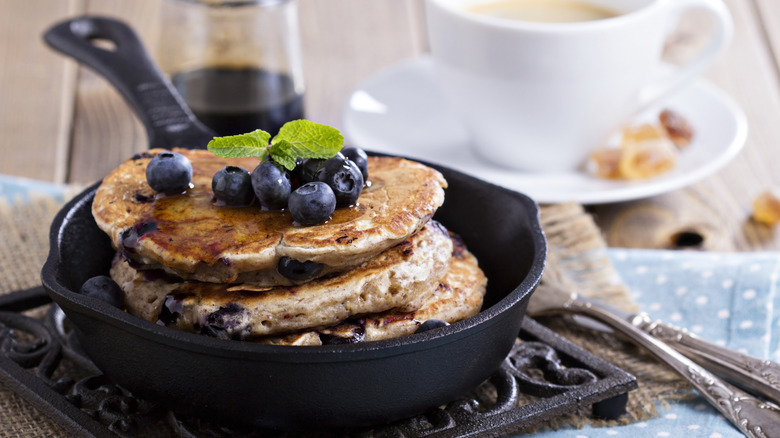 A stack of blueberry pancakes sit in a cast iron pan next to a cup of coffee and silverware