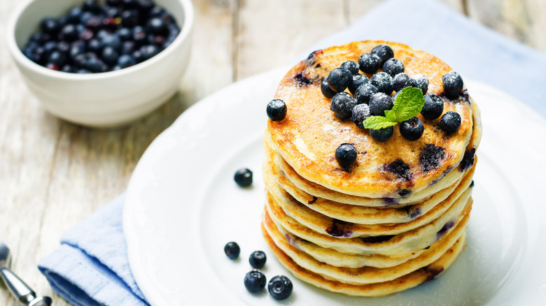 A stack of blueberry pancakes sit on a white plate next to a bowl of blueberries