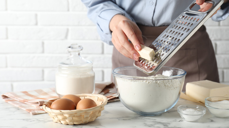 A person grates butter into a bowl of flour