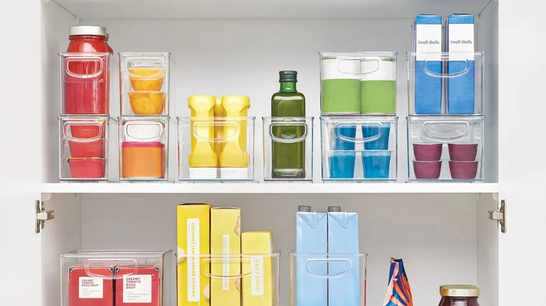Clear iDesign 18-piece kitchen and pantry bin set displayed with assorted snacks, condiments, and bottled drinks.