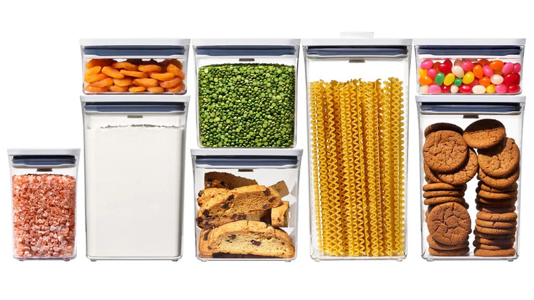 Eight clear OXO POP food storage containers with white push-button lids filled with dry goods like pasta, flour, and snacks.