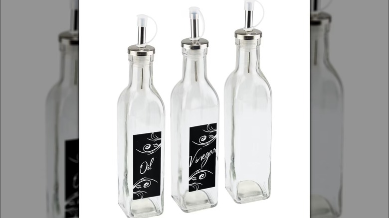 Three clear glass bottles with pour spouts, one with a decorative black "oil" label, one with a black "vinegar" label, and one with no label
