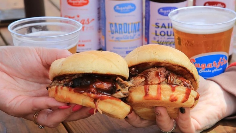Two hands holding burgers with Sugarfire sauces and beer in the background
