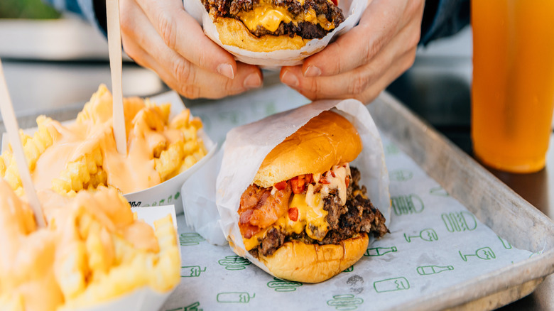Person in blue shirt holding cheeseburger in white wrapper above a tray of fries and a cheeseburger